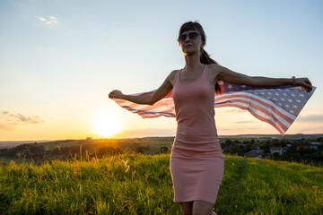 Back view of happy young woman posing with USA national flag outdoors at sunset. Positive girl celebrating United States independence day. International day of democracy concept.