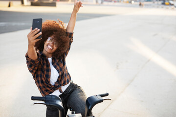 Young lady on electric bicycle on the street. Beautiful african girl taking selfie photo