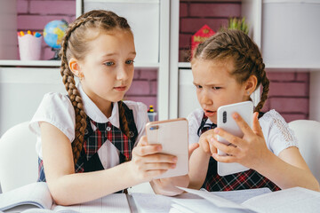 Two young female classmates are sitting at a desk and using smartphones. Modern technology concept.