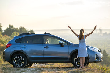 Happy young woman driver with outstretched up hands enjoying warm summer evening standing beside her car. Travelling and vacation concept.