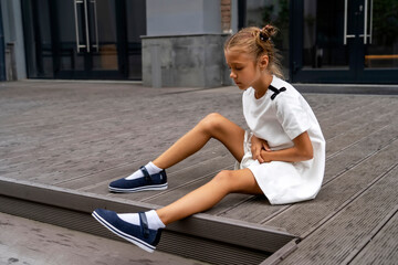 Fototapeta na wymiar Little girl in white dress sitting on steps on street has stomachache. Kid holds hands on belly and suffers. Stomach pain.