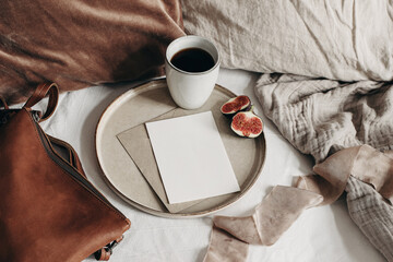 Autumn breakfast in bed composition. Blank greeting card mockup, cup of coffee and fig fruit on ceramic plate. White linen background. Velvet cushions and leather bag. Thanksgiving, fall. Top view. - obrazy, fototapety, plakaty