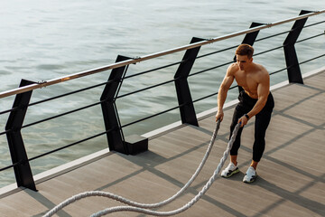 Obraz premium Strong man training with fighting ropes, Sportsman doing battle rope training outdoors on the sea on the pier