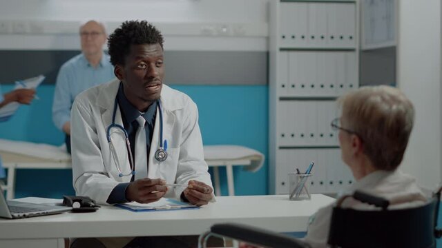 Black medic talking to disabled patient with handicap at desk in healthcare office. African american doctor helping elder woman with disability sitting in wheelchair. Old person at checkup