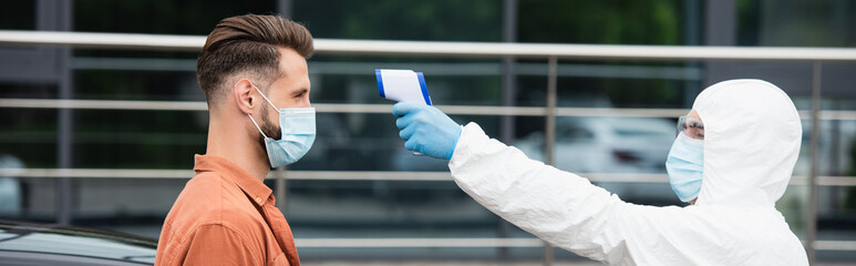 Medical worker holding pyrometer near driver in medical mask outdoors, banner