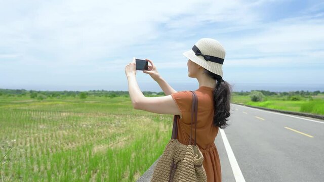 happy asian tourist is pivoting in one direction to take a panorama of the paddies with her smartphone on King Kong Avenue under blue sky in a rural area.