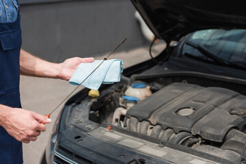 Cropped view of mechanic holding rag and dipstick while checking motor oil near blurred car