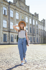 Fototapeta premium European educational or studying concept. Cheerful young blonde high school or college student female with backpack and laptop wearing jeans and hat standing in university campus. High quality image