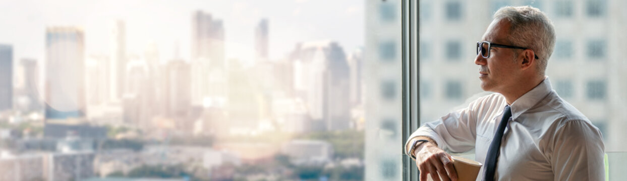 thoughtful senior business man holding cup of coffee standing over window glass in office on high building cityscape view background . worry old worker distracted . executive thinking and consider