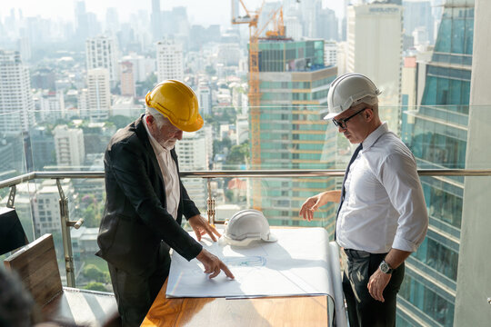 mature architect talking with senior business manager, Project owner with blueprint to construction on tall building cityscape background. old foreman discussion with contractor  builder on high site