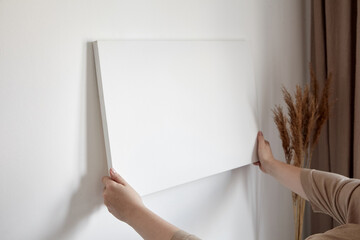 Blank canvas in female hands, picture mockup. Woman hanging canvas on white wall - obrazy, fototapety, plakaty