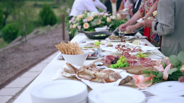 Decorated wedding buffet table with fresh flowers bouquets and variety of tasty italian prosciutto slices, cheese chops, fruits, snacks, canapes and fish appetizers. Wedding guests tasting traditional