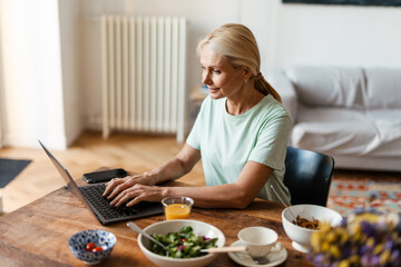 Obraz na płótnie Canvas Blonde mature woman using laptop while having lunch at home