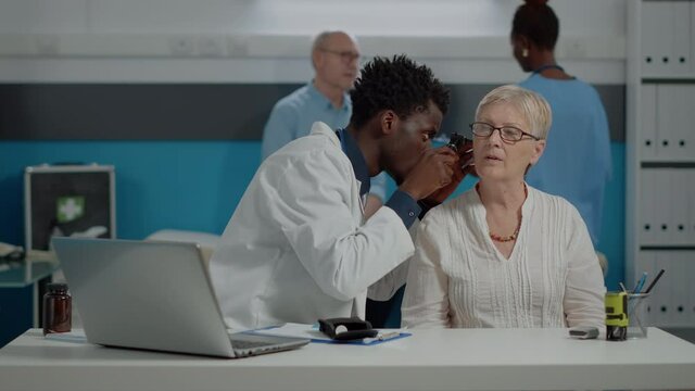Healthcare specialist holding professional otoscope for ear examination of elderly patient at desk in doctors office. Otologist using tool on caucasian senior woman with infection at clinic