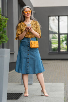 Mature Woman In Trendy Casual Clothes Using Smartphone Near Entrance Of Building