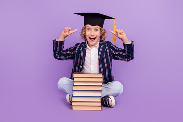 Portrait of cheerful schoolboy wearing demonstrating hat sitting pile book isolated over purple violet color background