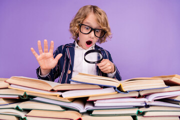 Portrait of trendy amazed schoolboy reading pile book using loop isolated over purple violet color background