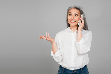 excited middle aged woman in white shirt gesturing while talking on smartphone isolated on grey