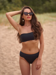 Attractive young sexy woman wearing black bikini swimwear standing and posing at sandy beach. Stunning girl with wavy brunette hair