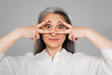 excited mature woman showing victory signs near eyes isolated on grey