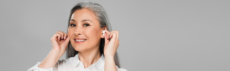 smiling middle aged woman inserting earphones while looking at camera isolated on grey, banner