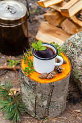 Obraz premium White campfire enamel mug with hot herbal tea on wooden stump. Bowler pot on background, cones, forest elements as decor. Concept of lunch break during hiking, trekking, active tourism, campring