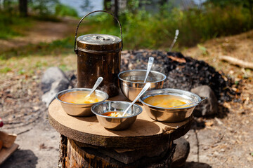 Lunch break at camp. Cooking simple delicious soup on fire, serving in touristic metal bowls. Concept of food preparation in travelling, camping, trekking, hiking trips. Bowler pot on background