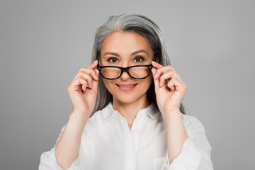 mature woman in white shirt taking off eyeglasses while smiling at camera isolated on grey