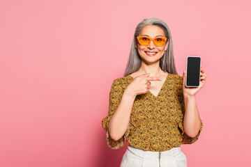 smiling woman in stylish sunglasses pointing at smartphone on pink background
