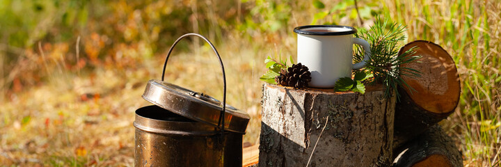 White campfire enamel mug with hot herbal tea on wooden stump. Bowler pot, backpack, cones, forest on background. Concept of lunch break during hiking. Active tourism, camping. Banner copy space