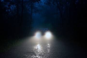 A spooky forest road with car headlights shining through the fog.