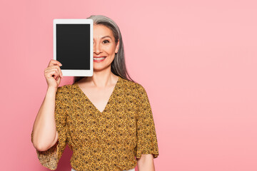 smiling middle aged woman obscuring face with digital tablet isolated on pink