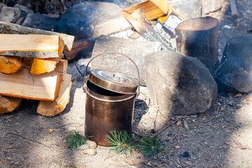 Metal bowler pot near bonfire and stake of wood. Preparation of soup or tea. Concept of traveling, active tourism, picnic cooking on fire. Close up