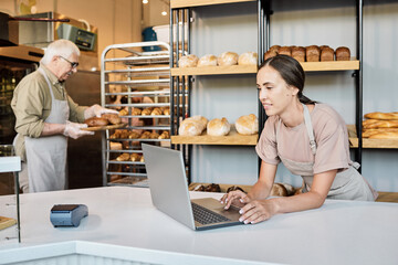 Young female baker using laptop against her mature male colleague carrying fresh bread