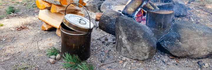 Metal bowler pot near bonfire and stake of wood. Preparation of soup or tea. Concept of traveling, active tourism, picnic cooking on fire. Close up banner