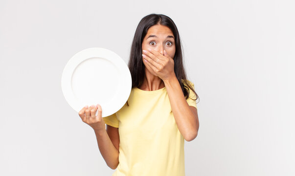 young hispanic woman covering mouth with hands with a shocked and holding an empty plate