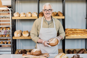 Happy aged baker with loaf of fresh bread looking at you by workplace - obrazy, fototapety, plakaty