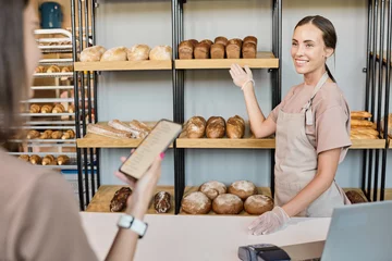 Tuinposter Young smiling clerk showing bakery assortment to female consumer by counter © pressmaster