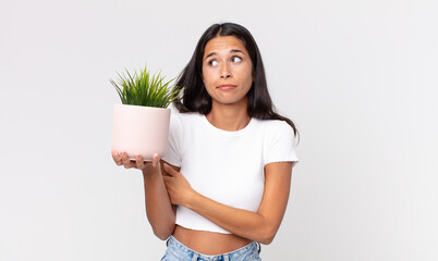 young hispanic woman shrugging, feeling confused and uncertain and holding a decorative house plant