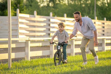 Happy young man helping his little son to ride bicycle along green lawn on sunny day