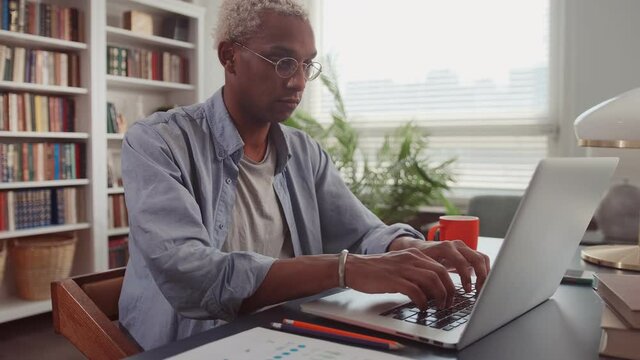 Inspired young African American male writer typing on laptop, writing article for online publication, visiting muse. Journalist working at home living room remotely via Internet. People and technology