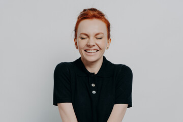 Happy pleased red haired woman with closed eyes and toothy smile posing against grey wall