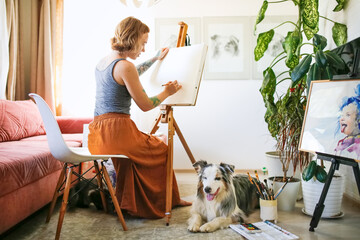 A cute red-haired girl artist draws together with the Aussie dog, Australian Shepherd breed. Owner and pet together in a bright living room, home workshop