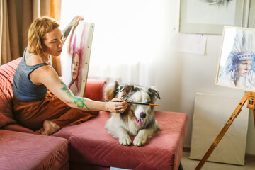 A cute red-haired girl artist paints together with the Aussie dog, the Australian Shepherd breed. Owner and pet together in a bright living room, home workshop, lifestyle in real room