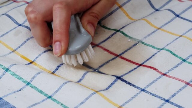 Rubbing wet stain with brush and washing white powder close-up, putting detergent on spoiled dirty on checkered towel. Concept of laundry, washing by hands. Stain remover. 