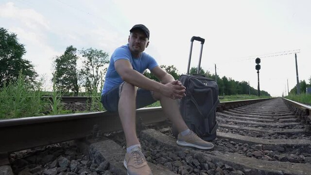sad man in blue shirt is late for train sitting on rails of railway looking on sides outdoor