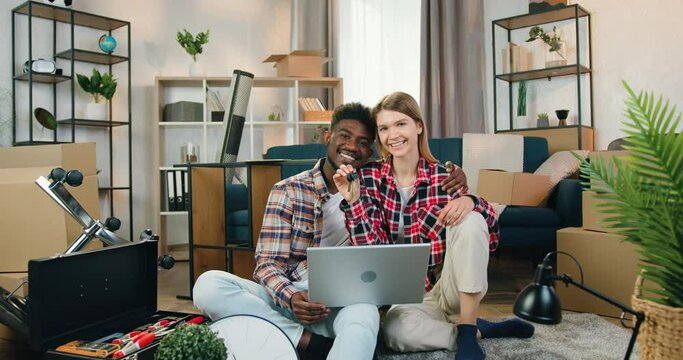 Good-tempered smiling happy mixed race couple sitting on the floor with huggs in not yet fully equipped room and showing keys from their new house into camera
