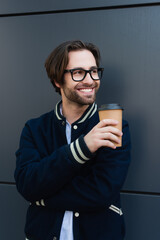 cheerful man with coffee to go looking away near grey wall outdoors