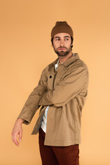 young man in jacket and brown beanie looking away while standing isolated on beige