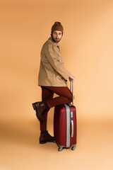 man in brown pants, boots and jacket standing near suitcase on beige